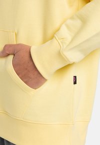Yellow sweatshirt made of soft material with a front pocket, ribbed cuffs, and a small black label with pink logo detail on the side.