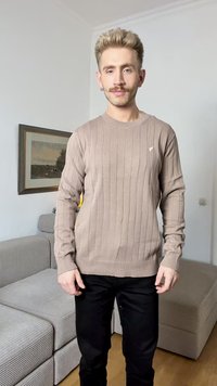Maglione beige a costine con maniche lunghe, scollatura rotonda e un sottile logo sul petto. Indossato con pantaloni neri. Tessuto morbido e design aderente.