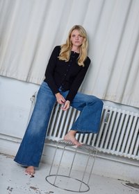 Cardigan nero con bottoni, jeans blu a gamba larga e piedi nudi, seduto su uno sgabello di filo metallico davanti a uno sfondo di tende bianche e termosifone.