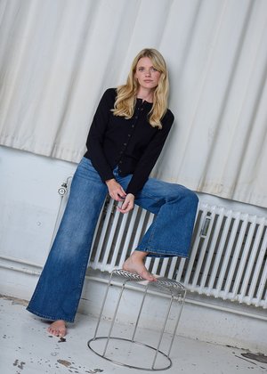 Melna pogāžama kardigāna, plašu zilu džinsu un basām kājām, sēž uz vadu krēsla pret balto aizkaru un radiatora fona.