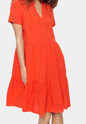 Frau trägt ein hellorangefarbenes, mehrlagiges Kurzarmkleid mit V-Ausschnitt vor einem einfarbigen Hintergrund.