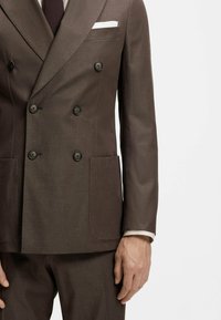 Veste de costume marron croisée avec boutons noirs et pochette blanche, portée avec un pantalon assorti et une cravate foncée.