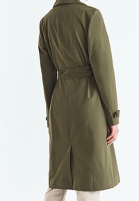 Long manteau trench vert olive avec taille ceinturée, poignets boutonnés et texture lisse. Comprend un col classique et une coupe jusqu'aux genoux.