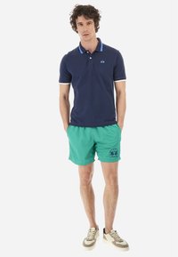 Marinblå polo med blå krage och logotyp, ihop med gröna badshorts med tryckt mönster, kompletterad med sneakers.