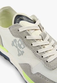 Baskets gris clair et blanches avec détails en daim, lacets blancs, logo tête de mort et os croisés sur le côté, et texte "Scalpers Company" sur la languette.