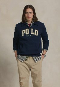 Mornarska mikica s logotipom "POLO", oblečena čez karirasto srajco. V kombinaciji z bež raztrganimi hlačami. Model stoji z rokami v žepih.