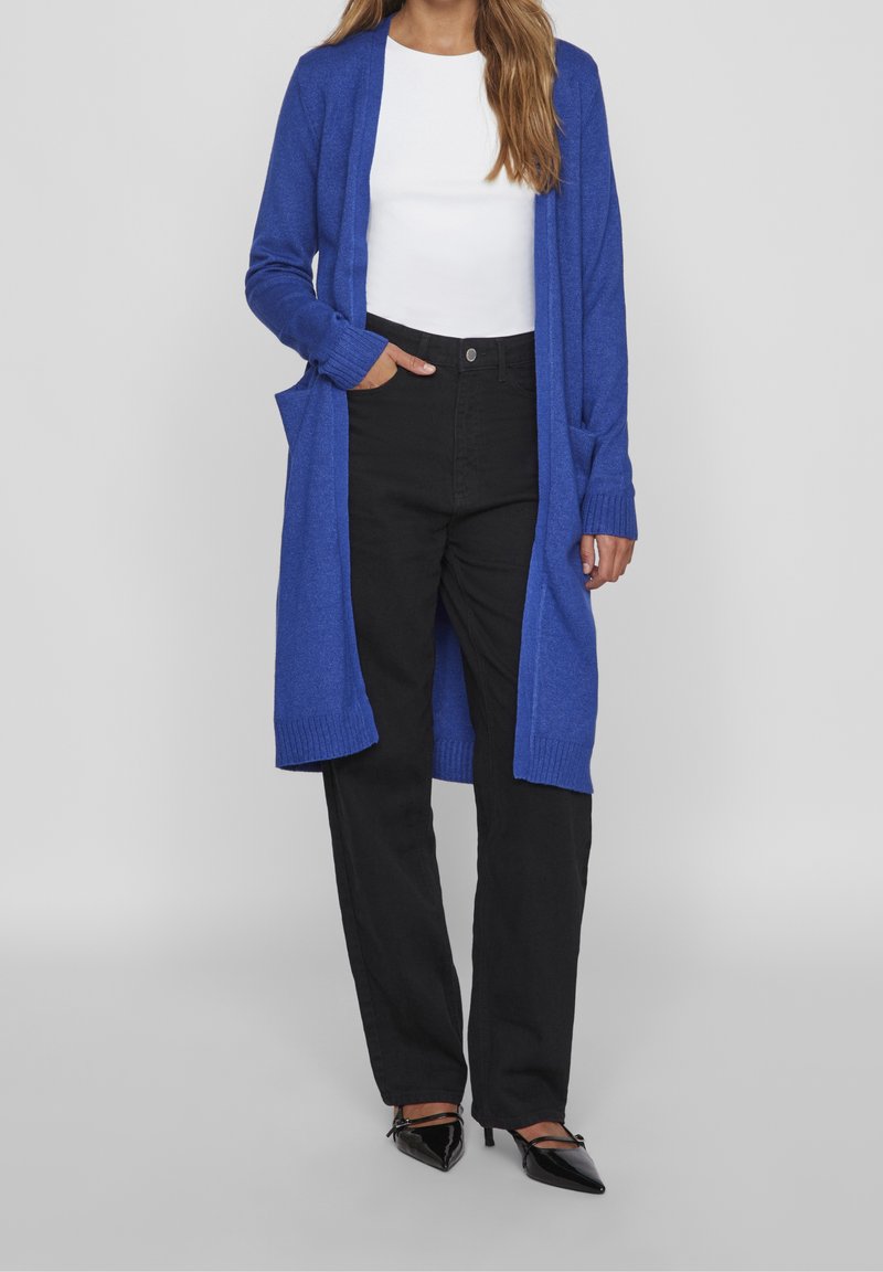 Femme portant un long cardigan bleu, un haut blanc, un pantalon noir taille haute et des escarpins noirs pointus, debout avec une main dans la poche.