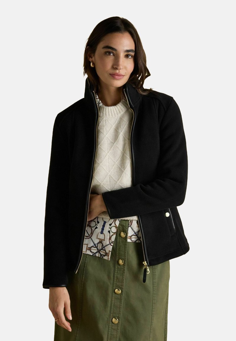 Žena oblečená v čiernej zipsovej bunde, smotanovom pletenom svetre, vzorovanej košeli a olivovo zelenej sukni na gombíky, stojí proti jednoduchému pozadiu.