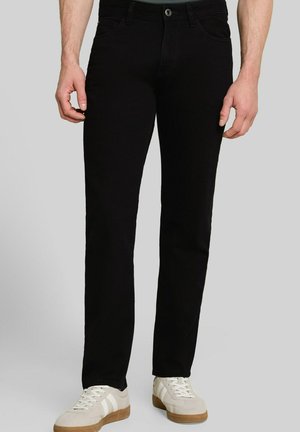 Homme portant un pantalon noir slim et des baskets blanches, debout devant un fond clair uni.