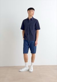 Marineblå kortærmet skjorte med knapper og brystlomme, parret med blå denim shorts og hvide sneakers, stående på en neutral baggrund.