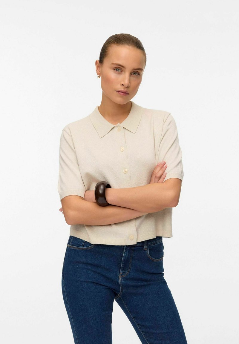 Kortärmad, ljusbeige stickad topp med krage och knappstängning. Matchad med blå jeans och ett mörkt armband på handleden.