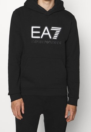 Mann trägt schwarzen Kapuzenpullover mit weißem und grauem "EA7 Emporio Armani" Logo und passende schwarze Hose.
