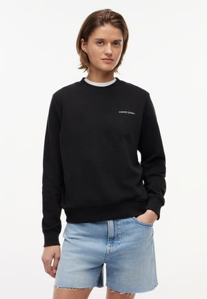 Mladý človek s krátkymi hnedými vlasmi oblečený v čiernej mikine "tommy jeans" a svetlomodrých rifľových šortkách, stojaci s jednou rukou v vrecku.