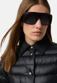 Schwarze Steppjacke mit hohem Kragen und silbernen Metallknöpfen, kombiniert mit großen schwarzen Sonnenbrillen mit dunklen Gläsern. Glatte Textur.