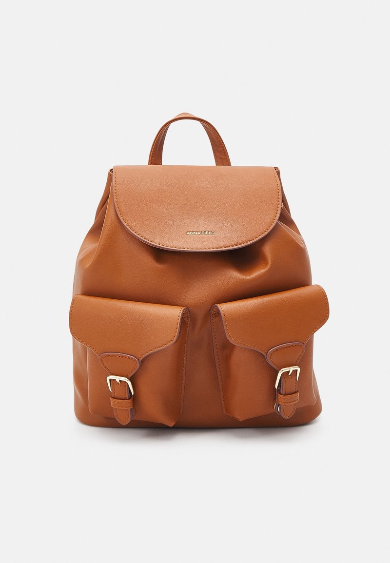 Mochila de cuero marrón con cierre de solapa, dos bolsillos frontales con hebillas y asa superior. Textura suave y detalles mínimos en el herraje.