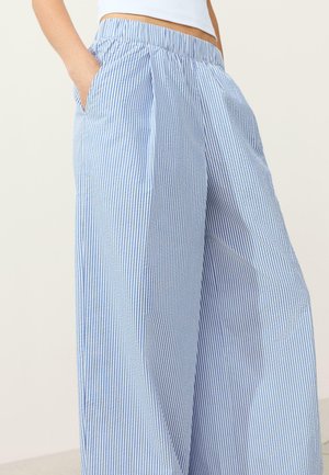 Žena nosiaca voľné, s vysokým pásom modro-biele vertikálne pruhované nohavice s rukami v vreckách, v kombinácii s bielym krátkym topom.