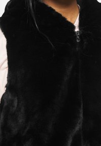 Musta tekoturkistakki, jossa on korkea kaulus ja vetoketju, ominaisuuksina pehmeä tekstuuri ja sileä pinta. Hienovarainen kiilto korostaa muotoilun yksityiskohtia.