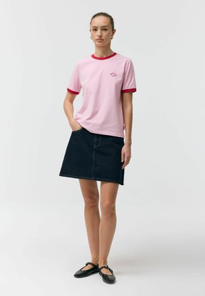 Kobieta stojąca z jedną ręką w kieszeni, ubrana w różowy t-shirt z czerwonym obszyciem, ciemną dżinsową spódnicę i czarne baleriny typu Mary Jane.