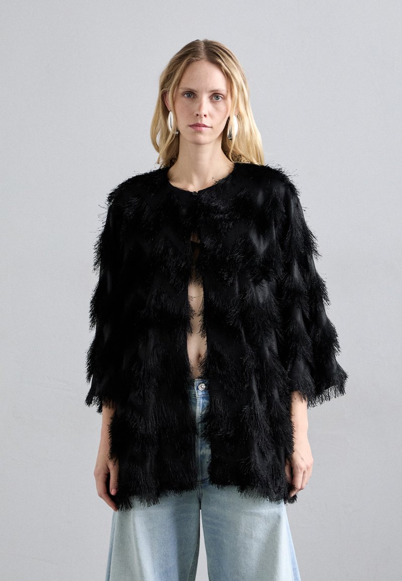 Melna, strukturēta jaka ar spalvām līdzīgu audumu, atvērta priekšpuse un platām piedurknēm, valkāta virs gaiši ziliem augstās jostasvietas džinsiem.