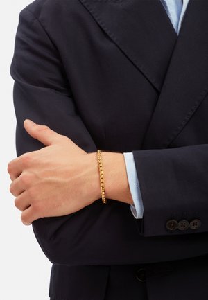 Goldene Kettenarmband, das an einem Handgelenk getragen wird und aus miteinander verbundenen Gliedern besteht. Die Hand ist auf einem Arm positioniert, der von einem dunklen Blazer bedeckt ist.