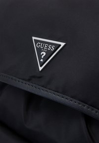 Bolsa de nylon negra con un parche triangular plateado que lleva el texto "GUESS USA" y un signo de interrogación. Textura suave y costuras resistentes.