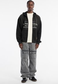 Schwarzer Kapuzenpullover mit Textgrafiken, über einem cremefarbenen T-Shirt getragen. Graue Cargo-Hosen mit Fronttaschen und schwarzen Sneakers.