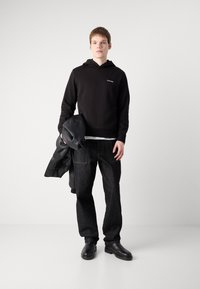 Svart hettegenser med logo, svart skinnjakke hengende over armen, mørke denimjeans og svarte sko, som står på en nøytral bakgrunn.