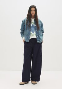 Giubbotto in denim, felpa bianca con grafica blu, pantaloni cargo blu scuro, scarpe slip-on in camoscio. Vestibilità rilassata, stile casual, con dettagli di cucitura visibili.