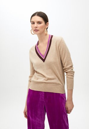 Maglione beige a V con rifiniture a strisce bordeaux e viola, abbinato a pantaloni in velluto rosa lucido. Il maglione presenta maniche lunghe e polsini a coste.