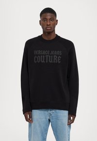 Černý svetr s raglánovými rukávy, který se pyšní texturovaným, embosovaným logem "VERSACE JEANS COUTURE" v tmavě šedé barvě. Nosit s modrými džíny.