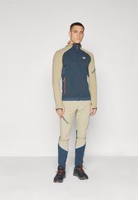 Men's activewear set featuring a navy and beige zip-up jacket with orange accents, paired with beige pants, and black footwear.