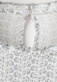 Biely textil s modrými vírivými vzormi, detailmi so zberaním a volánovým lemom pri výstrihu. Disponuje zapínaním na gombíky vzadu.