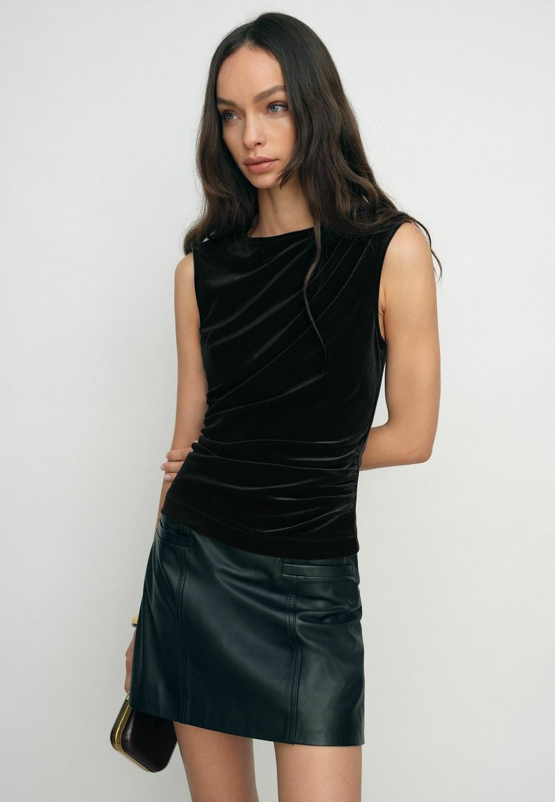 Čierny sametový top bez rukávov s drapovanou textúrou, v kombinácii s tmavozelenou koženou mini sukňou. Modelka drží malú kabelku.