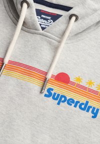 Felpa grigia con tasca a marsupio, caratterizzata da una superficie texturizzata, strisce orizzontali multicolori, una grafica del tramonto e il logo "Superdry".