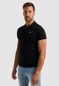 Polo noir avec une coupe ajustée, des manches courtes, une patte de boutonnage à trois boutons et une petite patch de marque sur la poitrine gauche. Porté avec un jean bleu clair.