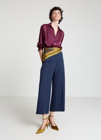 Chemise en satin bordeaux à boutons, pantalon large marine, écharpe à motifs dorés et bruns à la taille, et escarpins moutarde à talons aiguilles.