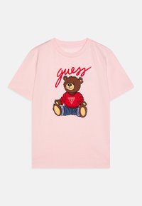 T-shirt rose à manches courtes avec un graphique d'un ours portant un pull rouge et un jean bleu, avec le texte "devine" en rouge au-dessus de l'ours.