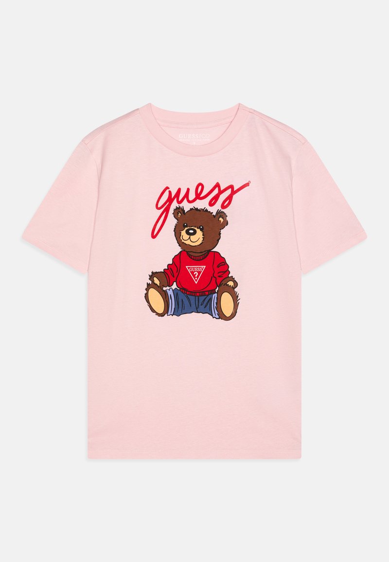 T-shirt rose à manches courtes avec un graphique d'un ours portant un pull rouge et un jean bleu, avec le texte "devine" en rouge au-dessus de l'ours.