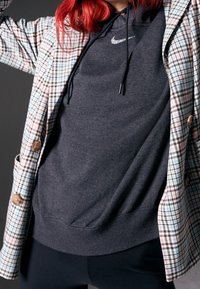 Chaqueta exterior de cuadros en colores pasteles con botones marrones sobre una sudadera gris oscuro con un logo de Nike plateado. Tela suave, corte relajado.