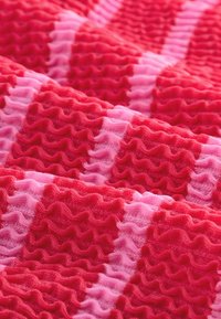 Gestructureerde stof in tinten rood en roze, met een golvend patroon van afwisselend gladde en geplooide delen, waardoor een ruitpatroon ontstaat.