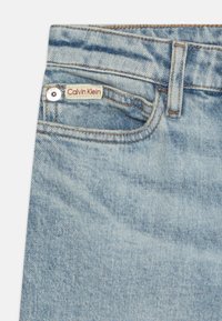 Svijetloplave traperice s klasičnim dizajnom od pet džepova. Imaju crveno-bijelu oznaku Calvin Klein na prednjem džepu s srebrnim detaljima.