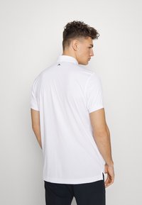Camisa polo branca com mangas curtas, colarinho e fendas laterais. A parte de trás exibe um discreto logotipo preto perto do pescoço.