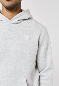 Bărbat purtând un hanorac gri deschis cu un mic logo alb brodat pe piept, vizibil de la gât până la talie, pe un fundal simplu.