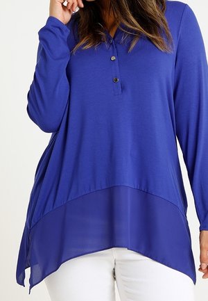Blouse bleu royal à manches longues avec col boutonné et ourlet asymétrique transparent, portée avec un pantalon blanc.