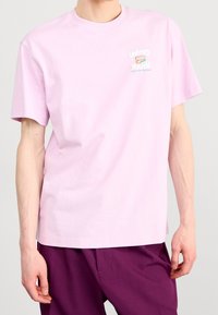 Osoba nosząca jasnoróżową koszulkę z krótkim rękawem z napisem i logo "United Youth" na piersi, w połączeniu z ciemnofioletowymi spodniami.