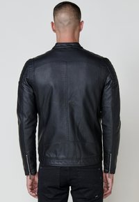 Homme à cheveux courts, de dos, portant une veste en cuir noir avec des poignets zippés et un pantalon noir, sur fond uni.
