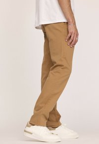 Pantalons en coton beige avec une coupe droite, dotés de poches latérales et d'une texture lisse, assortis à des baskets blanches et une chemise blanche simple.