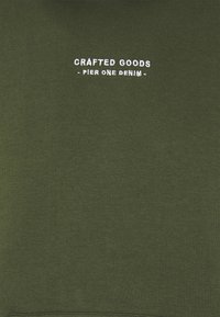 Sudadera verde oliva con texto bordado en blanco "CRAFTED GOODS - PIER ONE DENIM" en la parte delantera. Tejido de algodón suave, diseño minimalista.