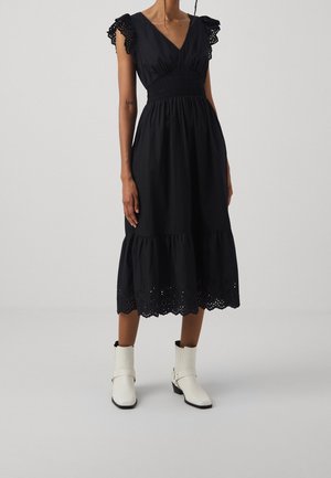 Kvinde iført en sort midi-kjole med flæseærmer og hulbroderet kant, kombineret med hvide ankelstøvler, stående mod en ensfarvet baggrund.