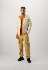 Beige puffer jacket, orange shirt, tan cargo pants with large pockets, and black shoes. Note: jacket has a hood and logo detail.